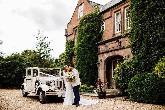 Bride and groom kissing in front of their wedding car outside Nunsmere Hall