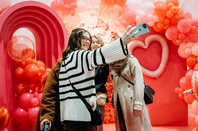 Group of women taking a selfie in front of a pink heart themed backdropped with balloons